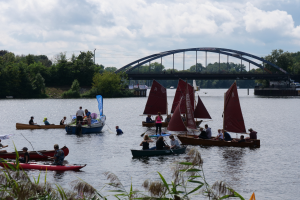 Eine Gruppe von Menschen in kleinen Booten auf einem Gewässer mit einer Brücke, Fahrzeugen, Bäumen und einem bewölkten Himmel im Hintergrund und Pflanzen am Boden, die ein Regatta suggerieren.