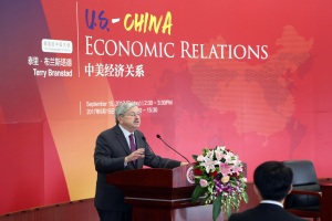 Ein Mann in einem schwarzen Blazer, weißem Hemd und lila Krawatte steht an einem Podium mit zwei Mikrofonen und einem Blumenstrauß, während er vor einer roten Wand mit einem Banner spricht, das "US-China Economic Relations" lautet. Ein weiterer Mensch sitzt rechts von ihm.