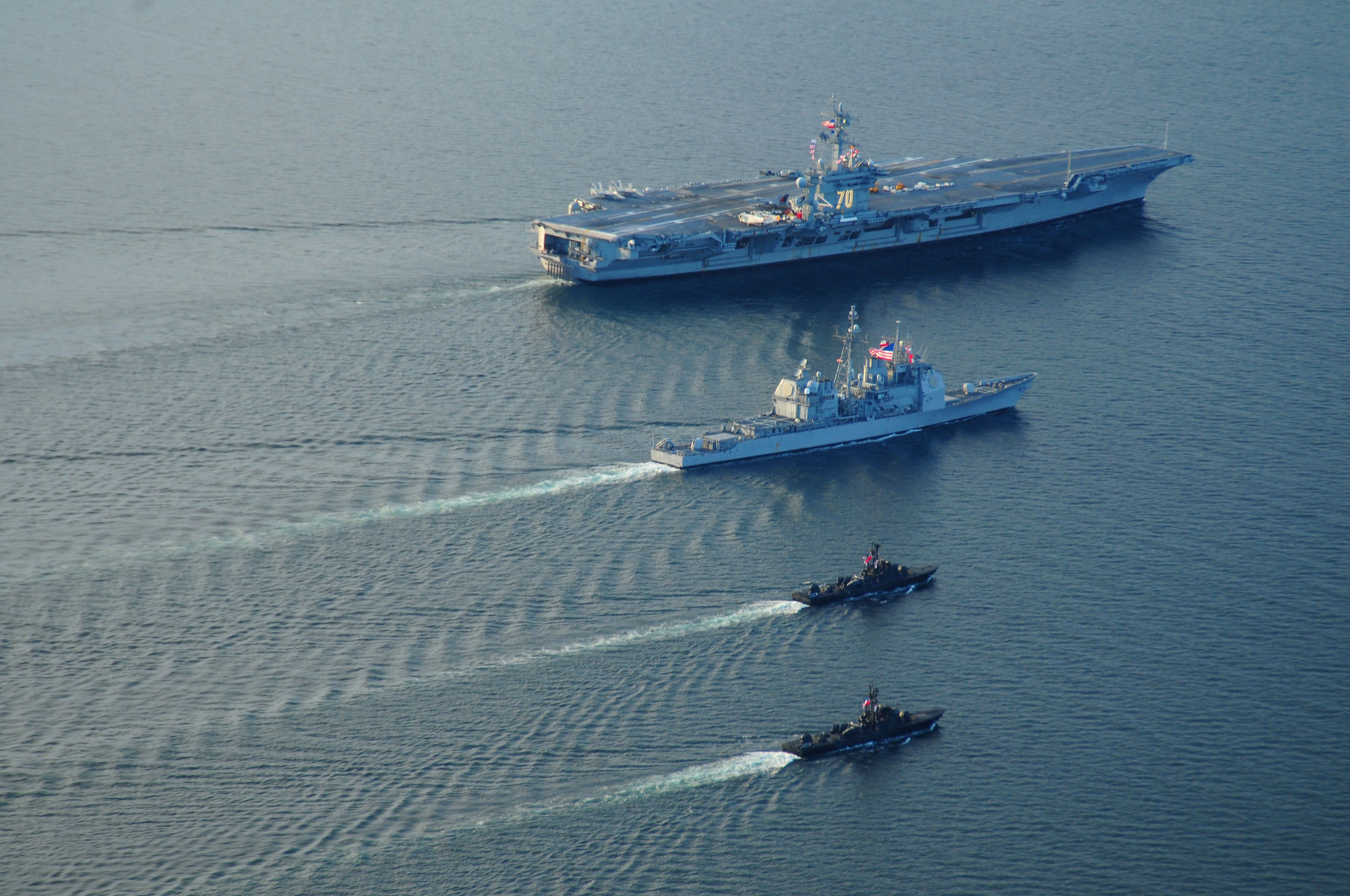 USS George H.W. Bush aircraft carrier (CVN-68) transiting the Strait of Hormuz with two small boats nearby, displaying flags and markings, and text at the bottom.