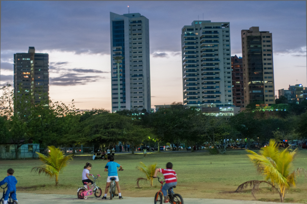 Kinder auf Fahrrädern in einem Park bei Sonnenuntergang, mit saftig grünem Gras und Bäumen, vor einem Himmel aus Orangen, Pinks und Purpurs, und im Hintergrund skylined Hochhäuser.