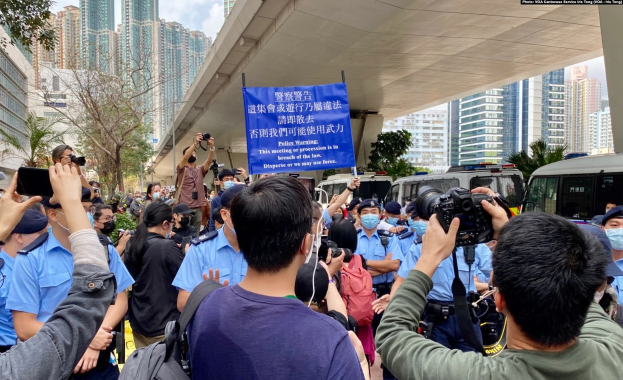 A group of people holding a banner reading "Hong Kong Police Wants to Stop the Violence Against Women" in front of a crowd, with vehicles, trees, buildings, and a cloudy sky in the background.