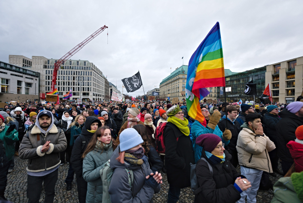 Eine große Gruppe von Menschen mit LGBTQ+-Rechten-Schildern und -Fahnen vor einem Gebäude mit einem Kran und einem bewölkten Himmel in Berlin.