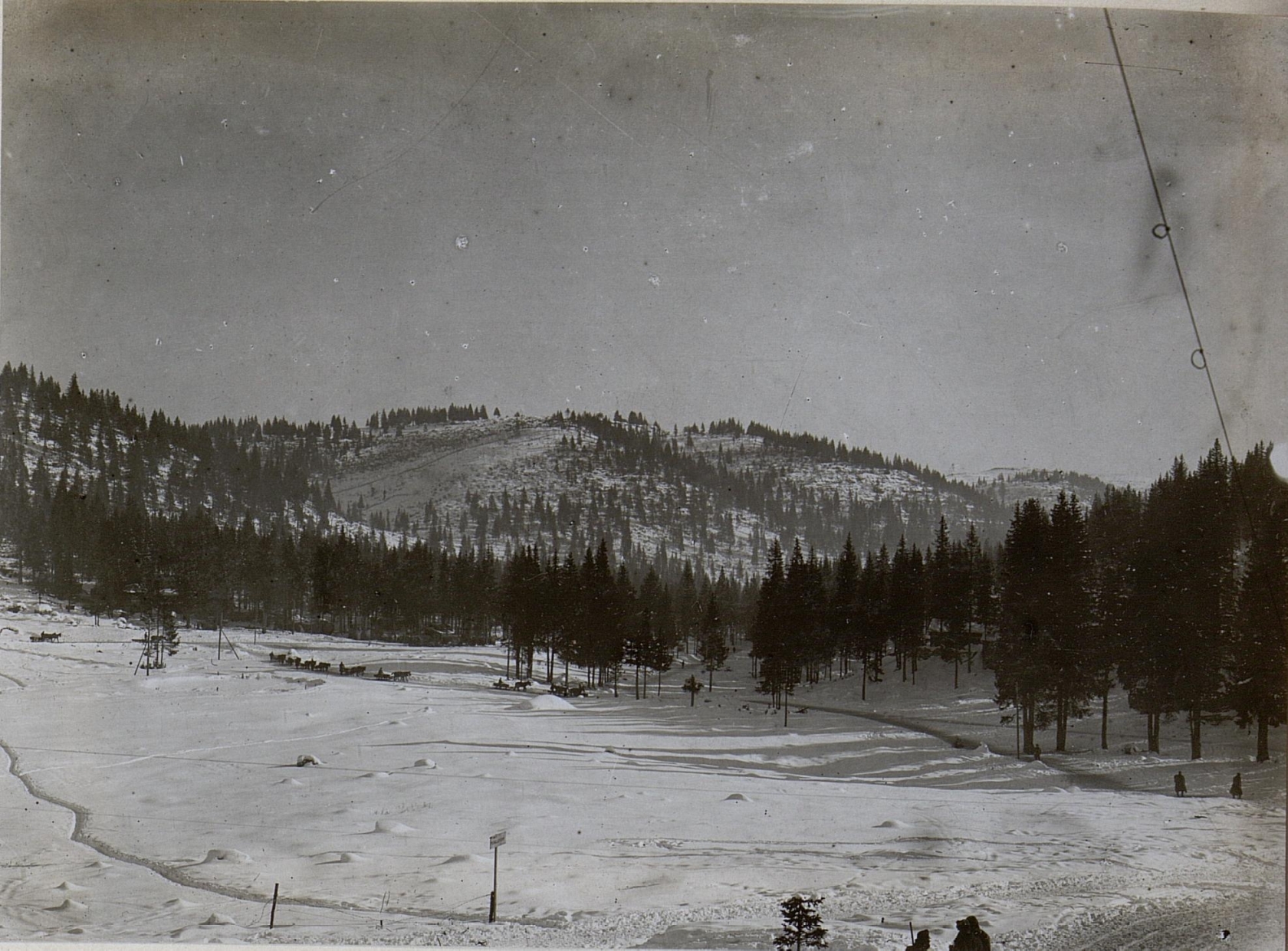 Ein Schwarz-Weiß-Foto einer Skipiste mit Menschen, die darauf Ski fahren, umgeben von Bäumen und Pfölen, mit Hügeln und einem klaren Himmel im Hintergrund und Text unten.