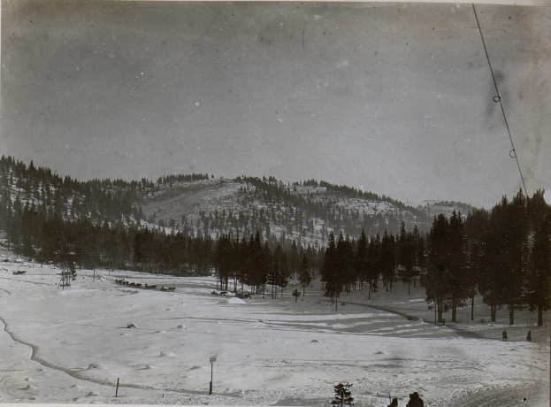 Ein Schwarz-Weiß-Foto einer Skipiste mit Menschen, die darauf Ski fahren, umgeben von Bäumen und Pfölen, mit Hügeln und einem klaren Himmel im Hintergrund und Text unten.