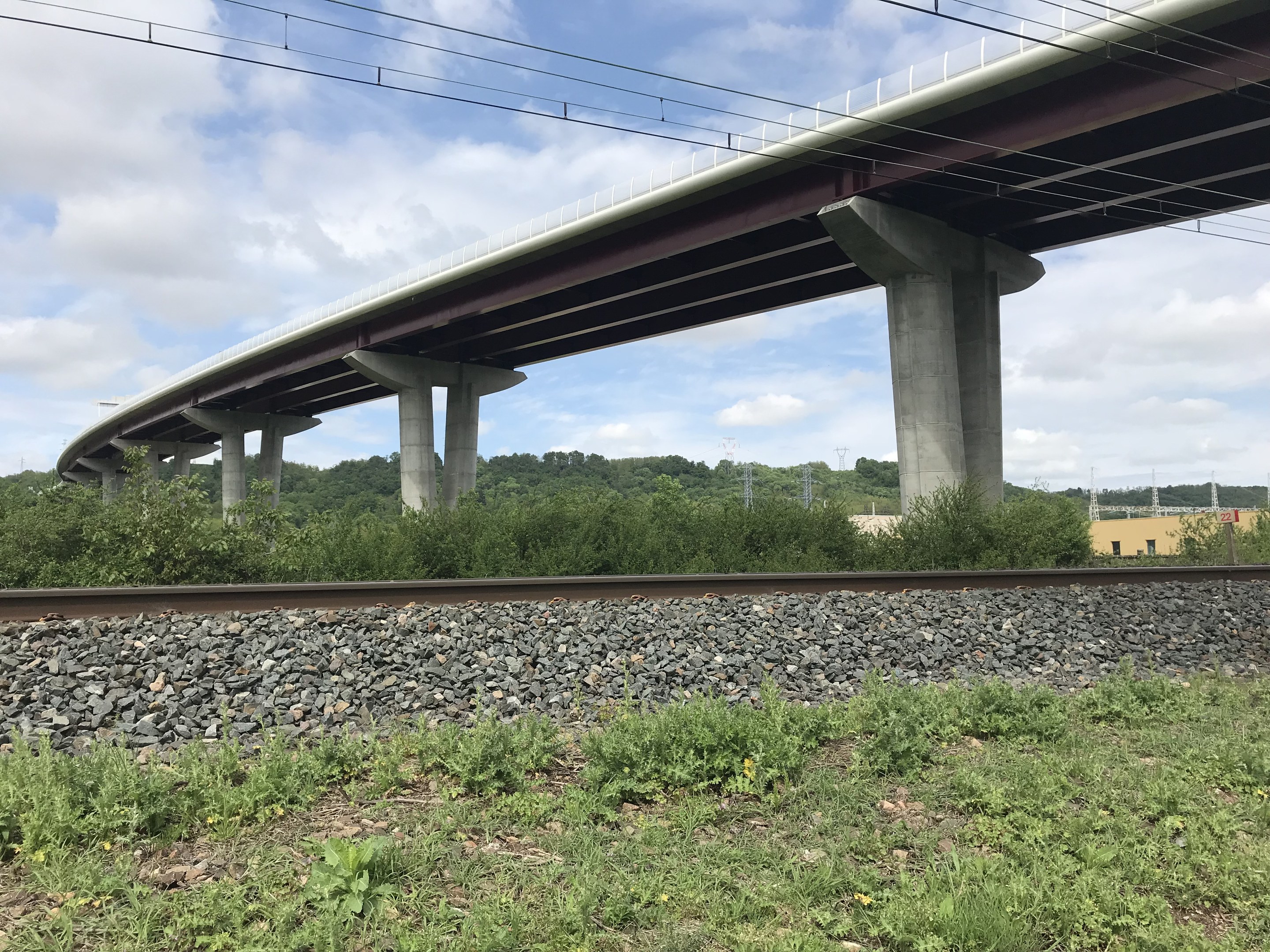 Eisenbahnschiene mit bewölktem Himmel, eine Brücke, die die Schiene überquert und renoviert wird, umgeben von Steinen, Gras, Pflanzen, Bäumen, Strommasten, Drähten und einem Gebäude im Hintergrund.