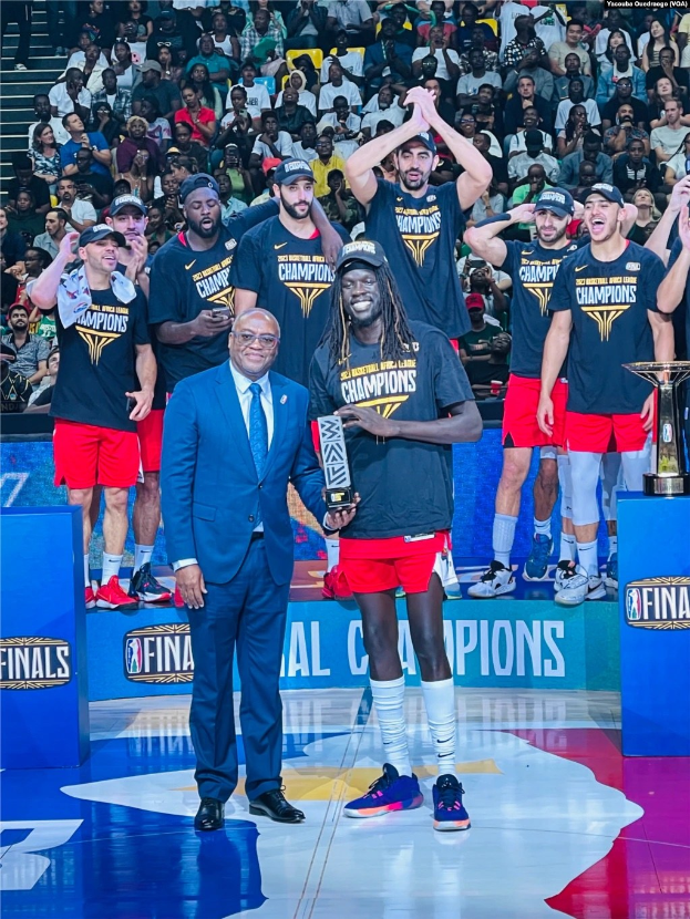 M├Ąnner feiern auf einem Basketballfeld mit Troph├Ąen in den H├Ąnden, mit zus├Ątzlichen Troph├Ąen auf Podesten im Hintergrund.