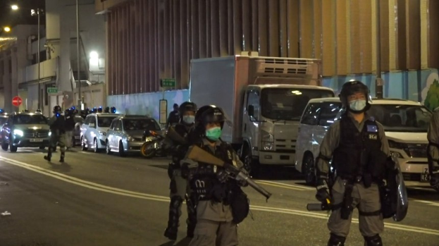 Eine Gruppe von Polizisten in Helmen, Handschuhen und Gesichtsmasken steht auf einer Stra├če bei Nacht, einige halten W├Ąffen, mit Fahrzeugen, Laternenmasten, Schildern und Geb├Ąuden im Hintergrund.