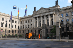 Large modern building with numerous windows, identified as the Bundestag in Berlin, Germany, surrounded by street infrastructure, vehicles, pedestrians, flags, and trees under a cloudy sky.