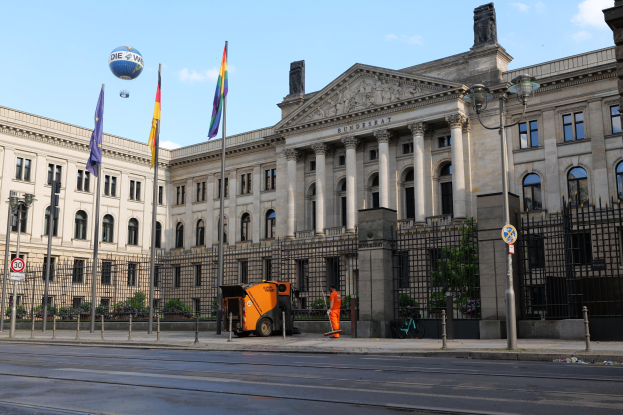 Large modern building with numerous windows, identified as the Bundestag in Berlin, Germany, surrounded by street infrastructure, vehicles, pedestrians, flags, and trees under a cloudy sky.
