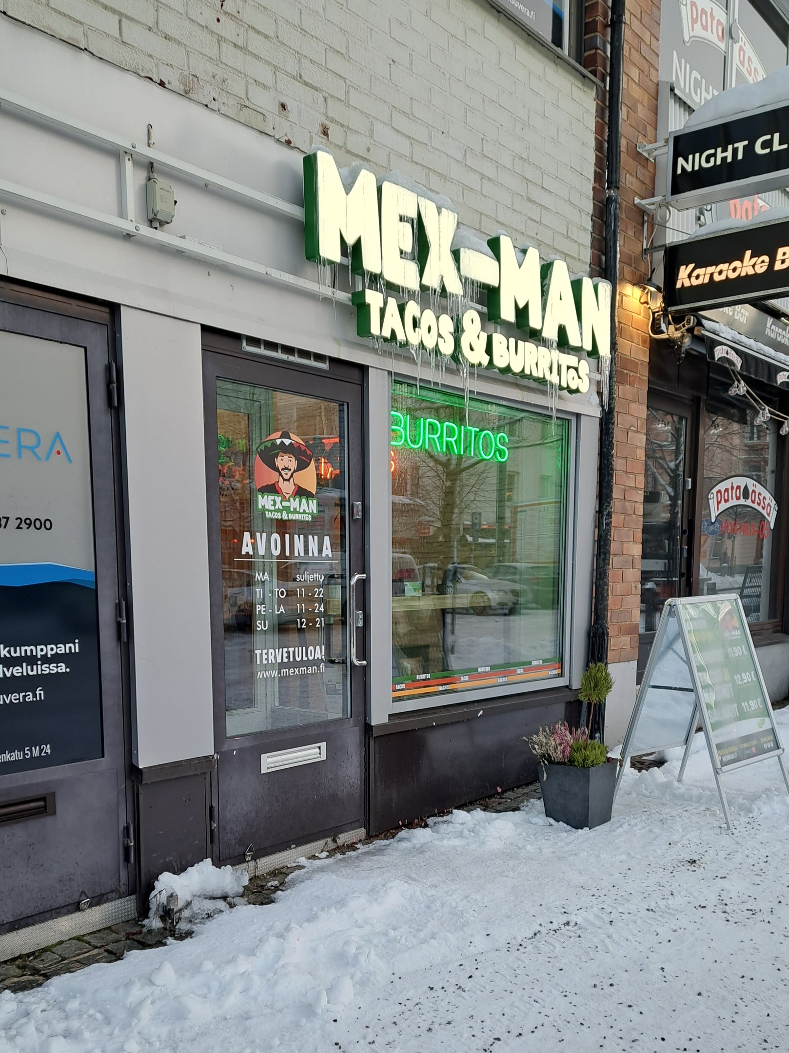 Ein mexikanisches Restaurant mit Glaswänden und -türen, das ein Schild mit der Aufschrift "mex-man tacos & burritos" sowie einen Topf mit einer Pflanze und Schnee auf dem Boden zeigt.