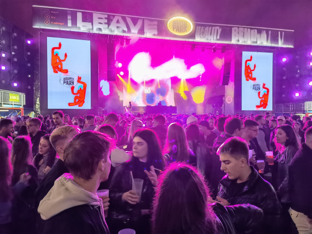 Eine große Menge steht vor einer hell erleuchteten Bühne auf einem Musikfestival, wobei einige Teilnehmer Gläser halten.
