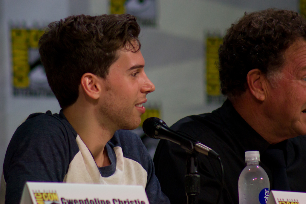 Ein Mann lächelt, während er vor einem Mikrofon steht, mit zwei Namensschildern und einer Flasche in der Nähe und einem Banner im Hintergrund.