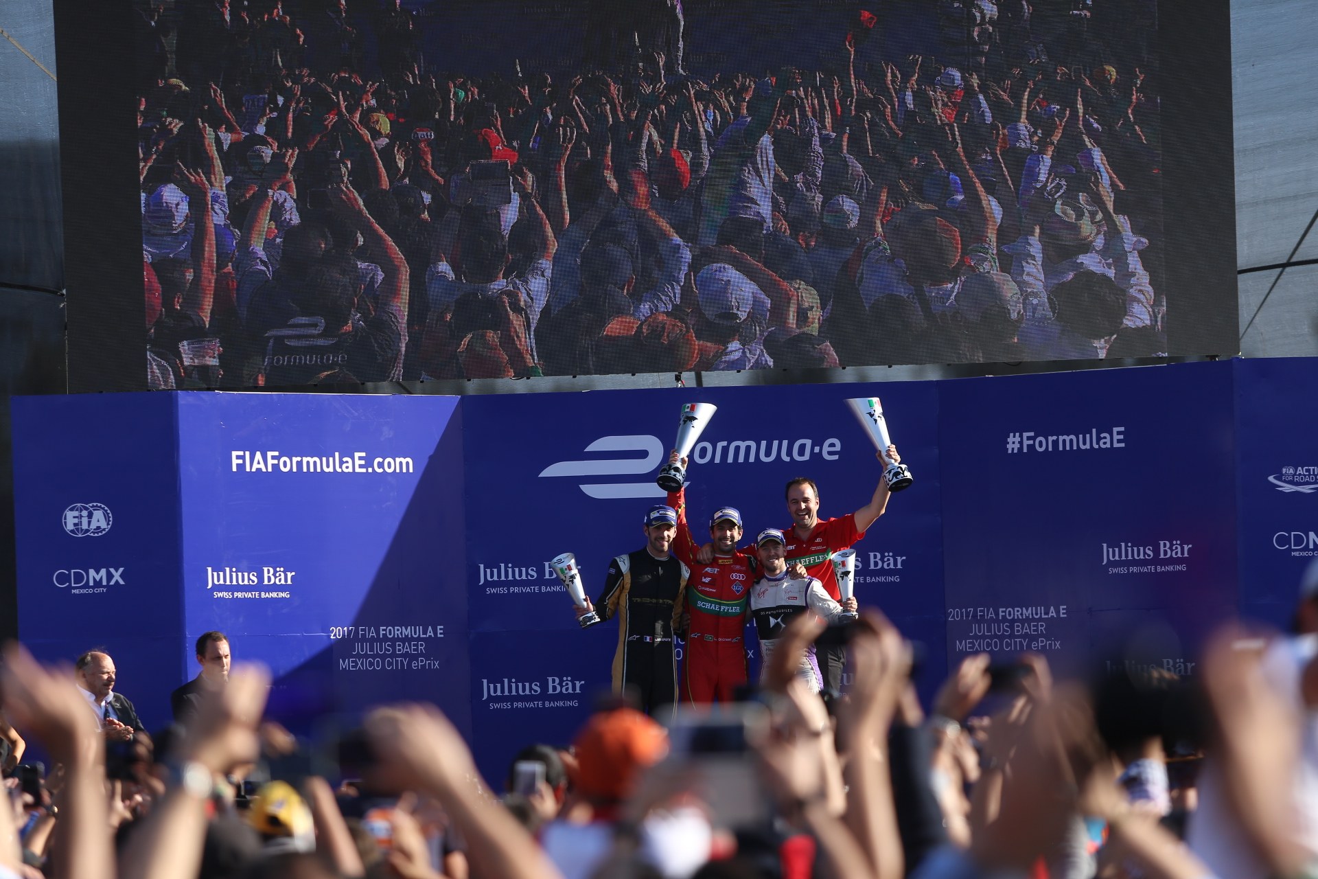 Gruppe von Menschen auf einem Formel-E-Podium in Mexiko-Stadt mit Tassen, Sponsorplakaten und einem Hintergrundbild.