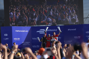 Gruppe von Menschen auf einem Formel-E-Podium in Mexiko-Stadt mit Tassen, Sponsorplakaten und einem Hintergrundbild.