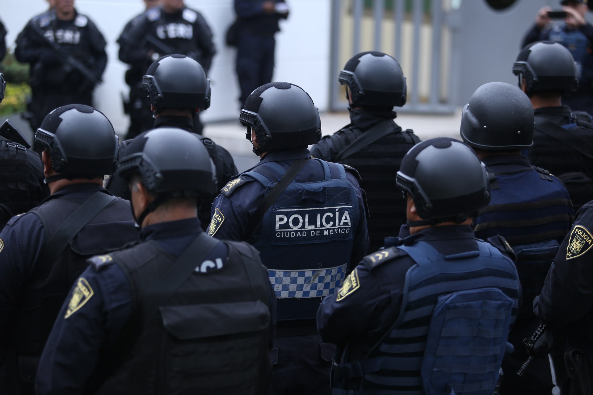 Mexikanische Polizeibeamte in Uniform und Helmen stehen vor einem Gebäude mit Menschen, einer Kamera, Pflanzen und einem Tor im Hintergrund.