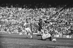Schwarzes und weißes Foto von Männern, die auf einem Feld Fußball spielen, mit Zuschauern im Hintergrund und einem Banner mit der Aufschrift "Bundesliga 1978-79 Ajax 1-1 Hannover 96."