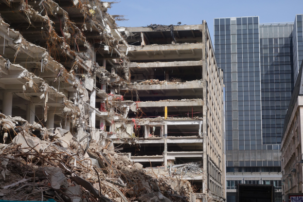 A large demolished building in an urban area, surrounded by intact buildings, street infrastructure, vehicles, trees, and a clear sky.