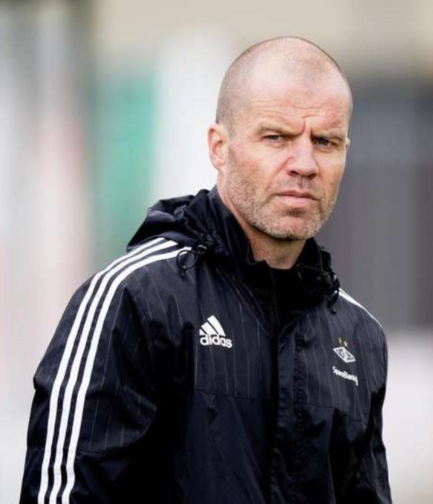 Ein kahlköpfiger Trainer in einer blauen Jacke mit weißen Streifen und einem weißen Kragen steht auf einem Fußballfeld mit verschränkten Händen und ernster Miene.