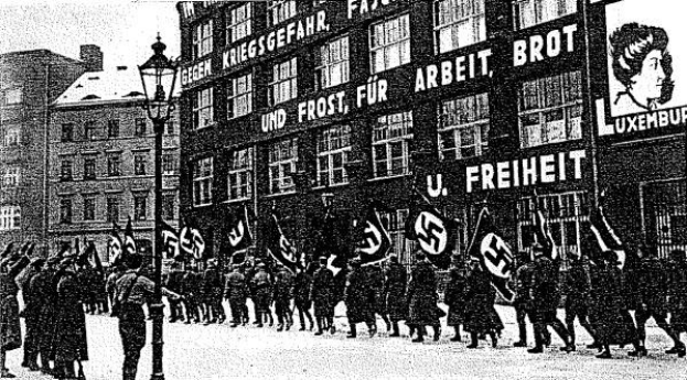 Black and white image of a group marching down a street holding flags and banners, with a light pole on the left and a building displaying a Nazi Party sign in the background.