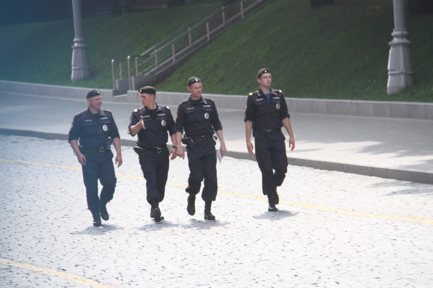 Drei Polizisten in Uniform gehen eine Straße entlang mit Pfählen, Geländern, Treppen und Gras im Hintergrund.