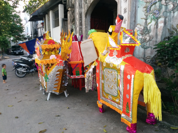 A vibrant Chinese New Year parade scene in Ho Chi Minh City, Vietnam, with vehicles, a person on the left, buildings and trees in the background, a wall painting on the right, and decorative parade items in the foreground.