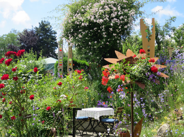 Ein Garten mit bunten Blumen und Pflanzen, der einen Tisch mit einer Decke und Stühlen zeigt, vor Bäumen und einem bewölkten Himmel auf der Chelsea Flower Show.