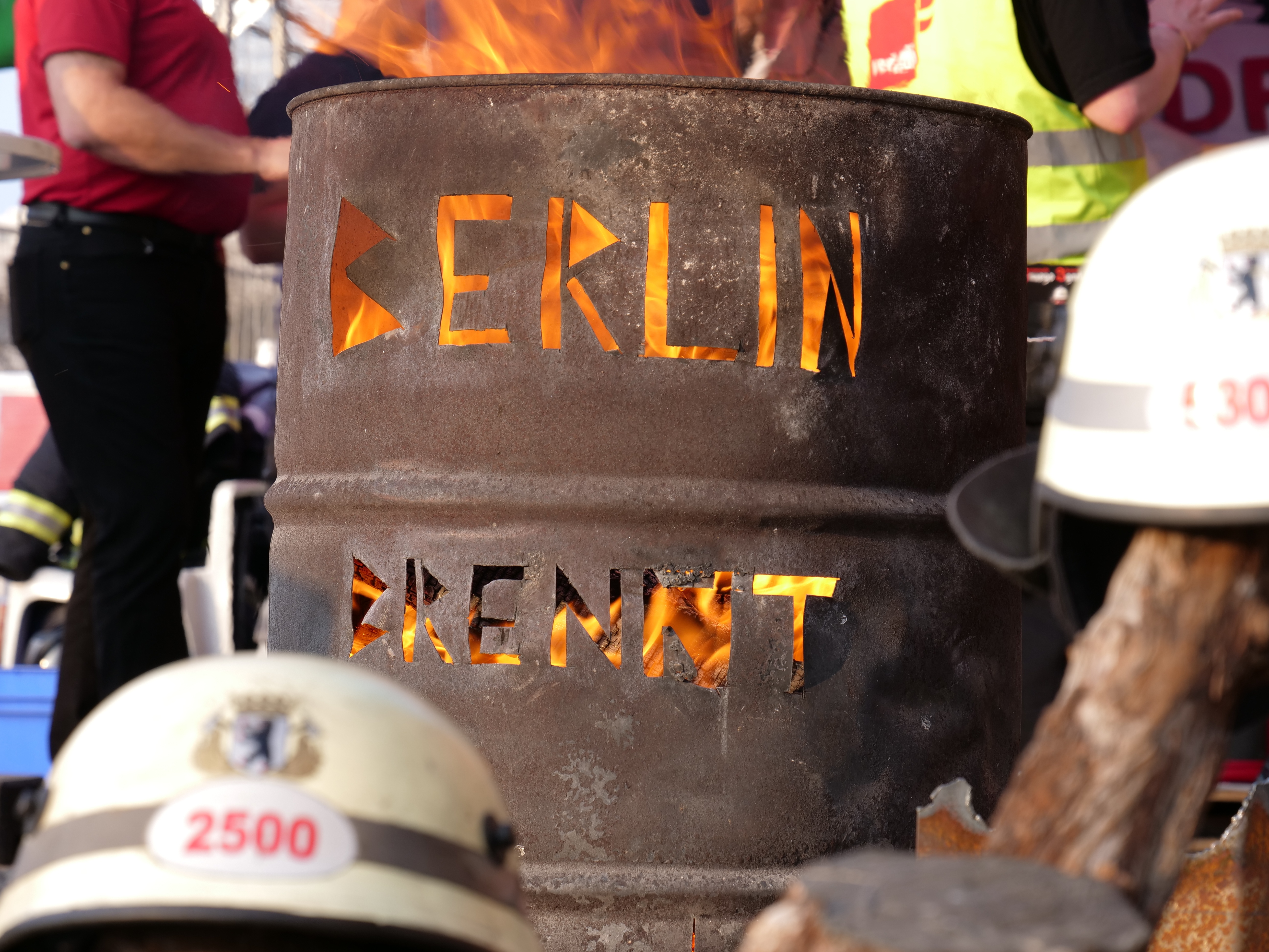 Eine Gruppe von Menschen um ein Feuerhydrant mit der Aufschrift 'Berlin' herumstehend, mit einem brennenden Fass im Vordergrund und Helmen unten und einem hölzernen Objekt auf der rechten Seite.