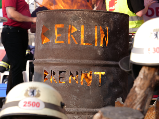 Eine Gruppe von Menschen um ein Feuerhydrant mit der Aufschrift 'Berlin' herumstehend, mit einem brennenden Fass im Vordergrund und Helmen unten und einem hölzernen Objekt auf der rechten Seite.