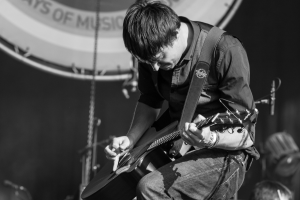 A man in a black shirt and jeans plays an electric guitar on a brightly lit stage, eyes closed in concentration, with an audience visible in the background.