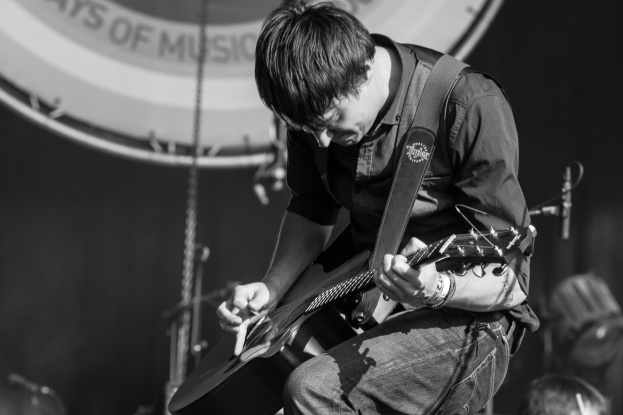 A man in a black shirt and jeans plays an electric guitar on a brightly lit stage, eyes closed in concentration, with an audience visible in the background.