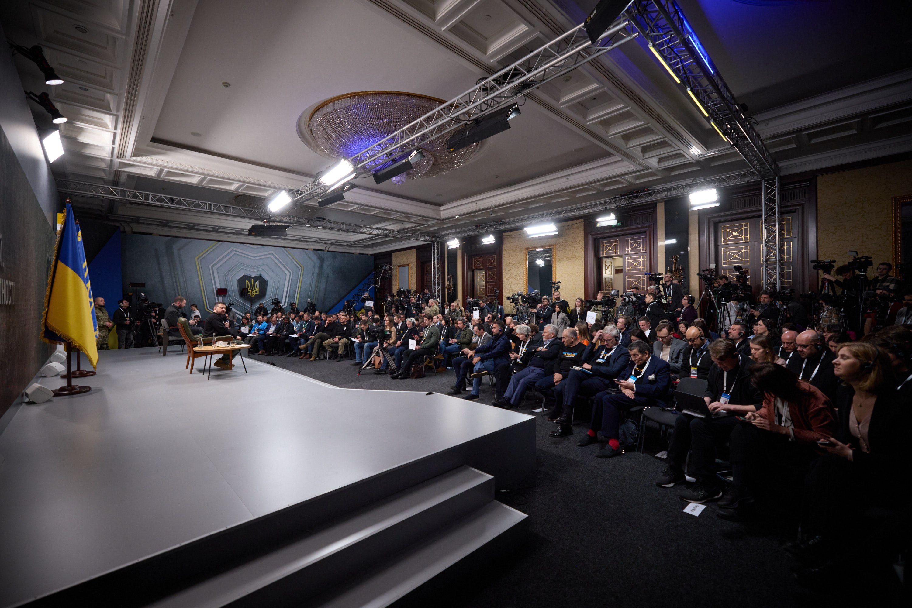 Eine grosse Gruppe von Menschen, die auf Stühlen vor einer Bühne mit einem zentralen Tisch sitzen, eine Flagge links und Fotografen rechts, was auf eine Pressekonferenz in der Ukraine hinweist.