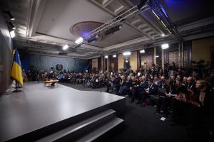 Eine grosse Gruppe von Menschen, die auf Stühlen vor einer Bühne mit einem zentralen Tisch sitzen, eine Flagge links und Fotografen rechts, was auf eine Pressekonferenz in der Ukraine hinweist.