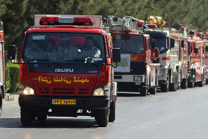 Eine Reihe von Feuerwehren mit Menschen darin fährt eine Straße entlang, flankiert von Pflanzen und Bäumen auf der linken Seite und mehr Bäumen im Hintergrund.