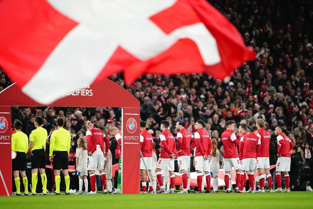Gruppe von Menschen auf einem Fussballfeld mit einer roten und weissen Flagge im Vordergrund, einem Bogen mit der Aufschrift "Bayern München vs Bayern München Wetten & Vorschau" im Hintergrund und einer grossen Menge im Stadion.