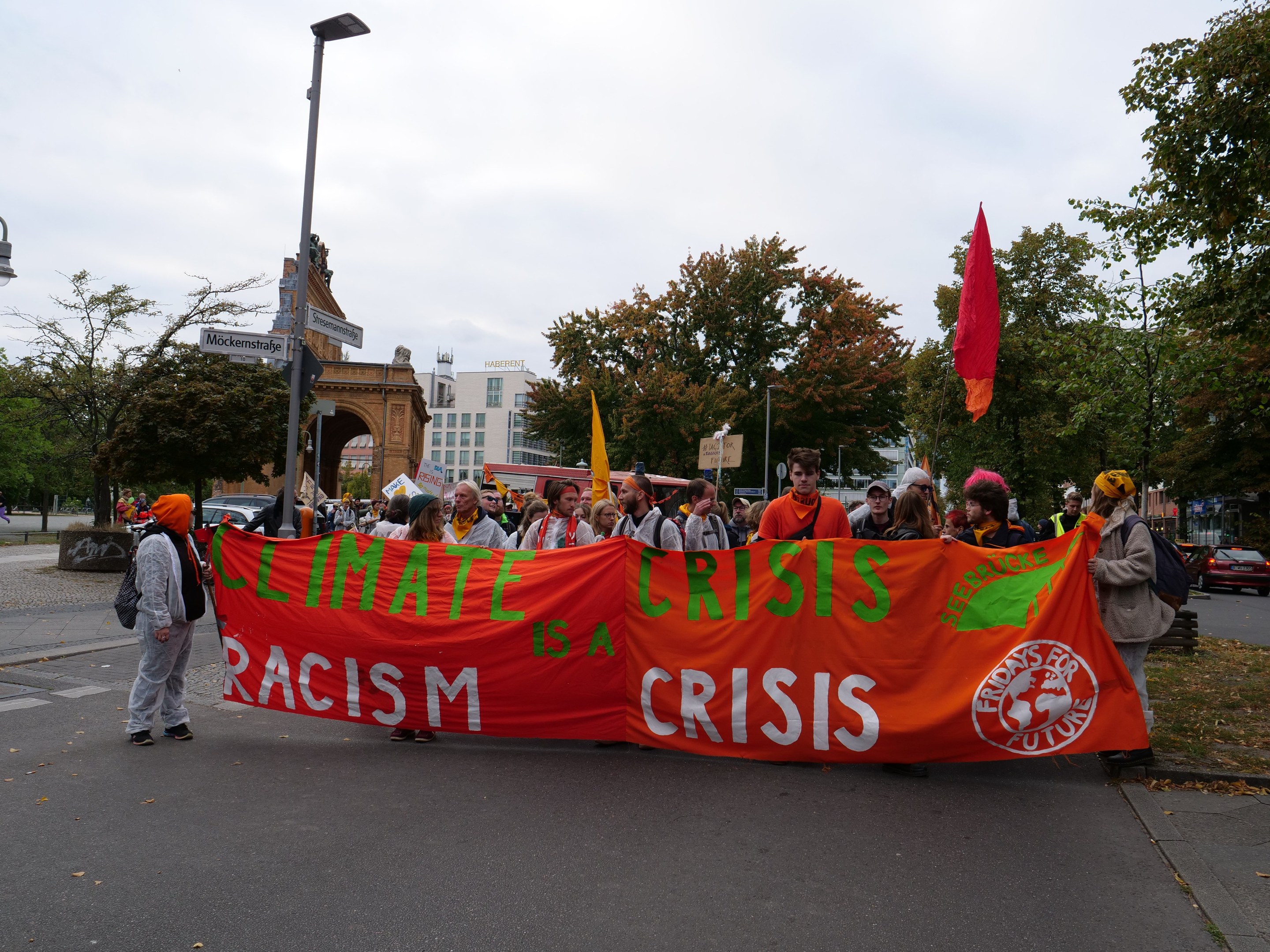 Eine Gruppe von Menschen marschiert eine baumbestandene Straße entlang und hält ein Banner hoch, auf dem "Klima-Krise ist eine Krise" steht, mit Fahrzeugen und Gebäuden im Hintergrund unter einem klaren blauen Himmel.