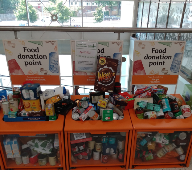 Orange Schränke mit Lebensmitteln wie Flaschen, Dosen und Kartons gefüllt, mit Informationsschildern und einem Blick auf Fahrzeuge, Bäume, Gebäude und Himmel durch Glasfenster.