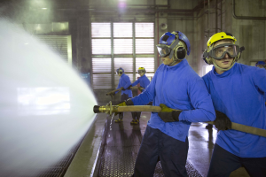 Eine Gruppe von Männern in blauen Hemden und gelben Helmen, die an Maschinen arbeiten, während einer das Boden mit Wasser besprüht, in einer Fabrik mit sichtbaren Rohren, Fenstern und Lampen.