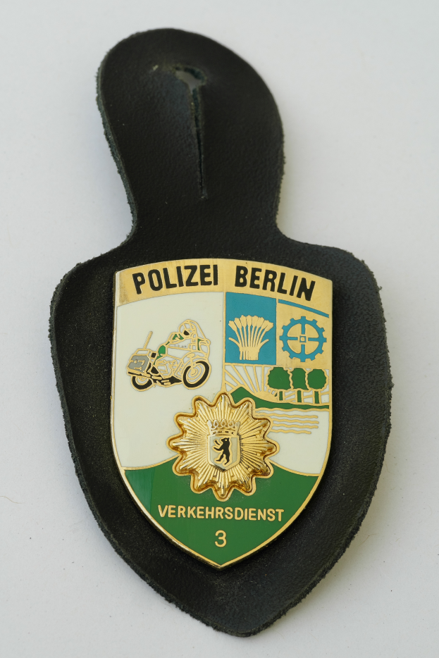 Ein Polizeistern mit der Aufschrift "Polizei Berlin" darauf, der ein blaues Schild mit einem weißen Stern in der Mitte zeigt, umgeben von einem weißen Rand, der auf einer Oberfläche liegt.