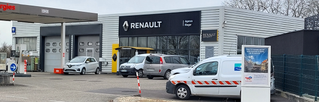 Ein Renault-Autohaus mit parkenden Autos vorne, umgeben von einem Metallzaun, einem Schild, Pfosten, einer Gruppe von Bäumen und einem bewölkten Himmel.