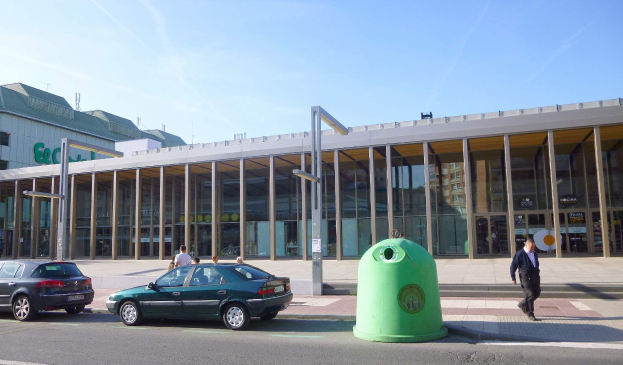 Grüner Müllcontainer auf einer Stadtstraße neben einem Glaswandgebäude mit Säulen, mit Fahrzeugen und Passanten in der Nähe und einer bewölkten Kulisse.