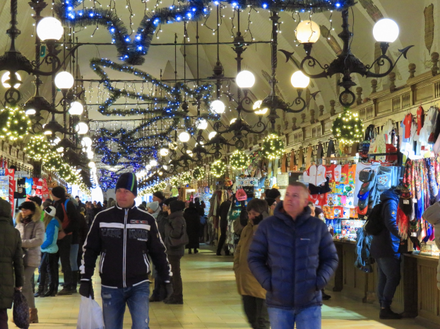 A bustling Christmas market with people walking, some wearing caps and holding bags, under hanging lights and decorations, with clothing and goods displayed on the right and a wall in the background.
