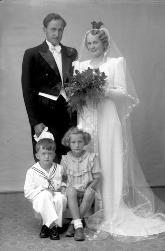 Schwarzes und weißes Foto einer Braut in einem weißen Kleid mit einem Strauß, einem grinsenden Bräutigam neben ihr und zwei Kindern, die vor ihnen auf dem Boden sitzen.