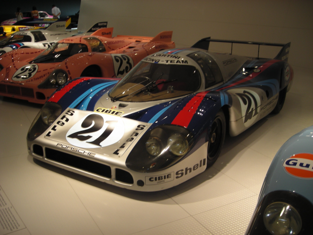 Ein Porsche 917K Rennwagen auf einer Museumsausstellung umgeben von anderen Sportwagen, mit Menschen in der Nähe und einer Wand im Hintergrund.