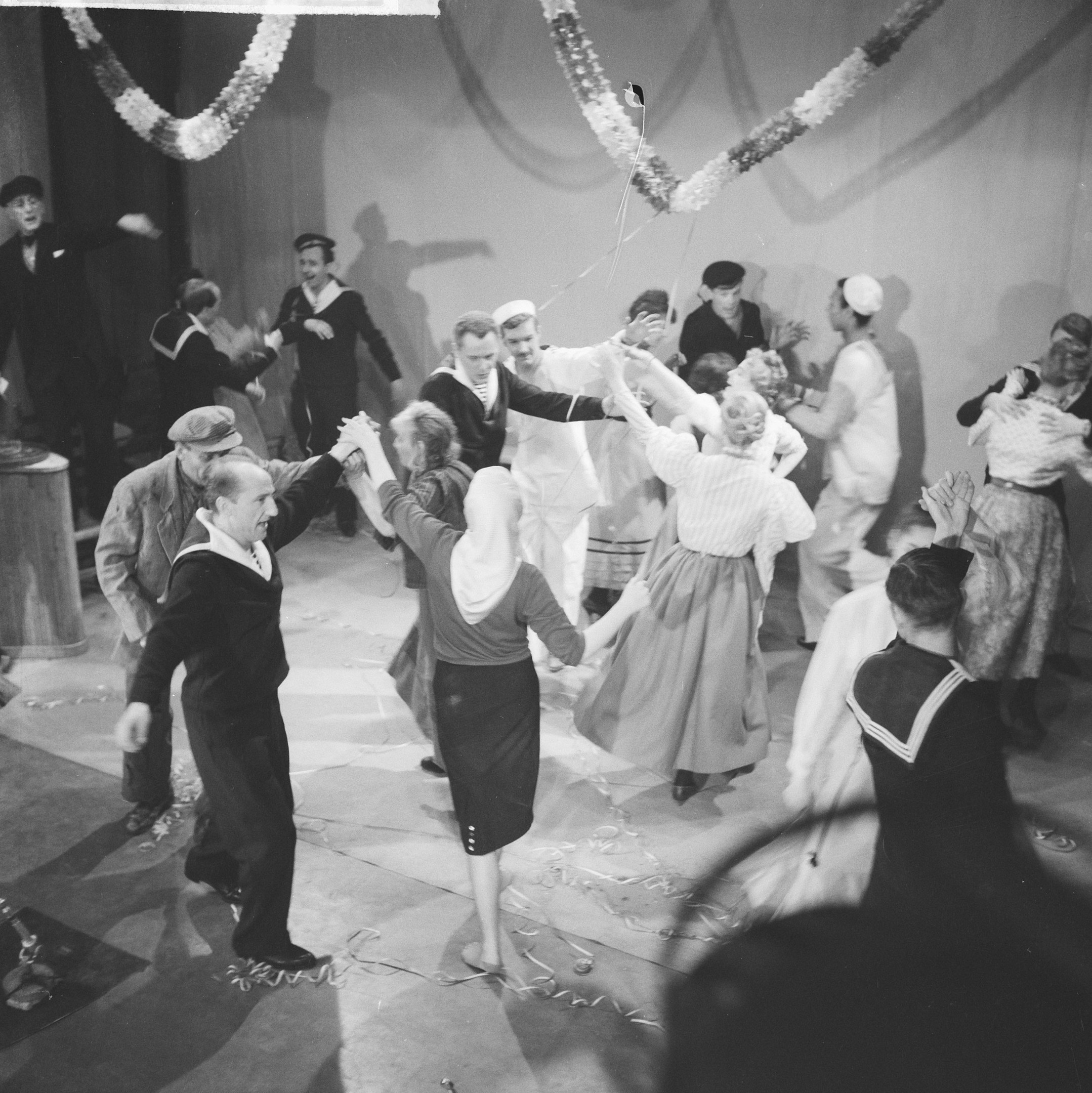 Schwarz-weißes Bild von Menschen, die auf einer Tanzfläche tanzen, mit einer dekorierten Wand dahinter und einem Tisch mit Gegenständen links.