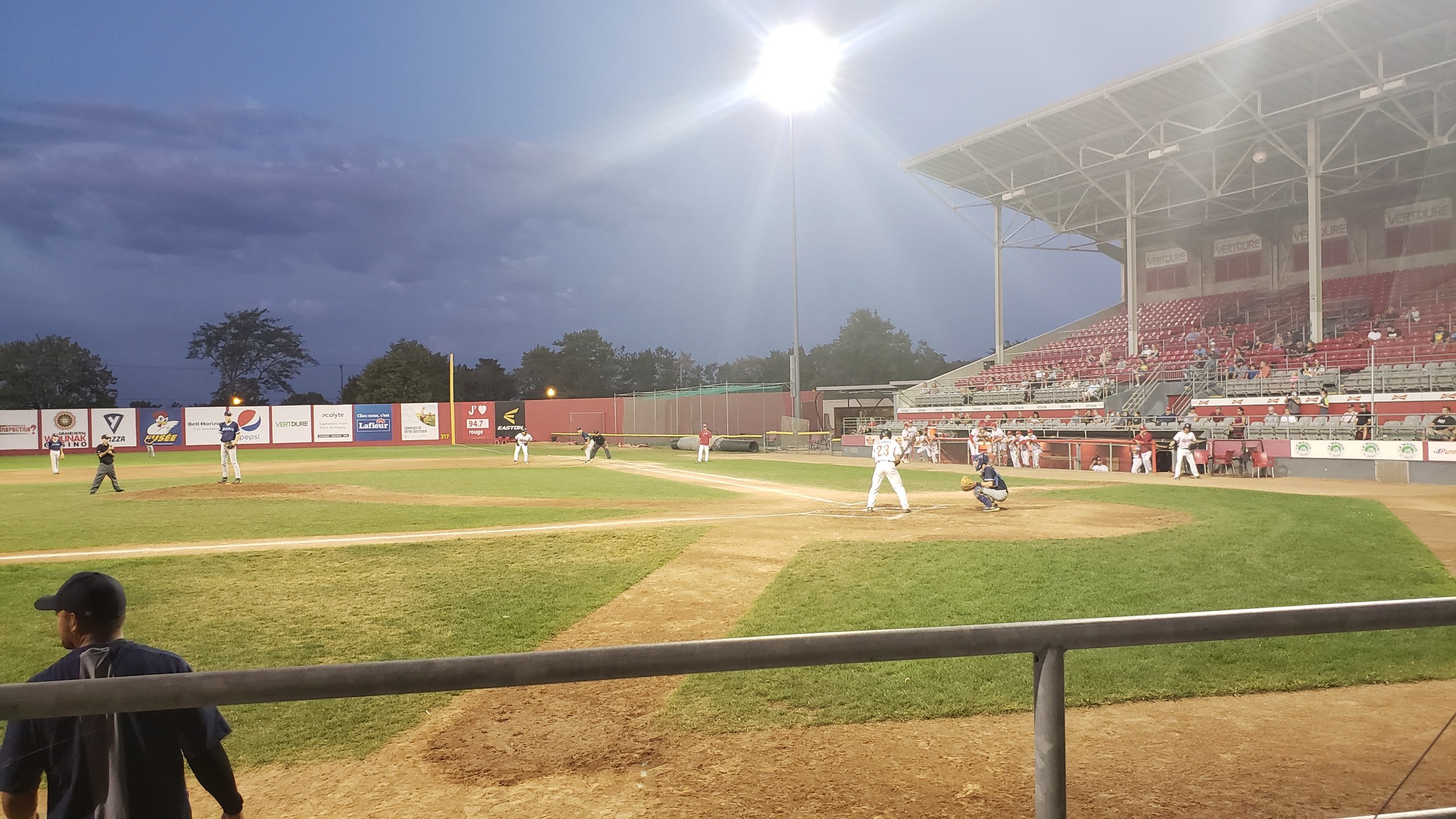 Baseballspiel in einem Stadion mit Zuschauern in den Rängen, Geländer im Vordergrund, Bäume, Mäste, Lichter, Werbetafeln und klarer blauer Himmel im Hintergrund.