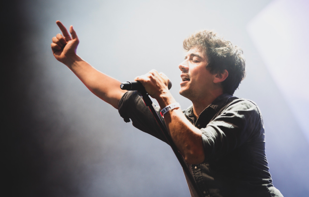 Ein Mann in schwarzem Hemd und Jeans singt in ein Mikrofon auf einer Bühne, mit hellen Lichtern und einem jubelnden Publikum im Hintergrund.