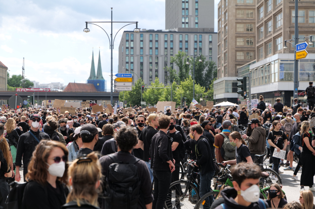 Eine große Gruppe von Menschen in Masken, die während einer Demonstration in Berlin eine von Bäumen gesäumte Straße entlanggehen, einige halten Fahrräder, mit Gebäuden, Verkehrszeichen und bewölktem Himmel im Hintergrund.