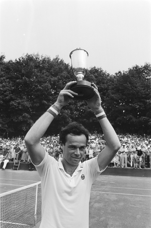Ein Mann auf einem Tennisplatz hält eine Trophäe in die Höhe, während ihn eine Menge hinter ihm anfeuert, mit Bäumen und einem blauen Himmel im Hintergrund.