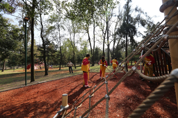 Gruppe von Menschen, die einen Seilparcours in einem Park mit Bäumen und Erde im Hintergrund absolvieren, mit einem Pfahl mit Seilen auf der rechten Seite.