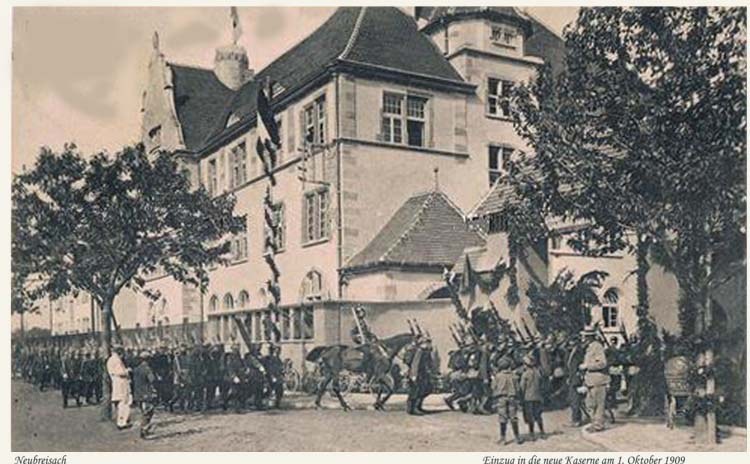 Black and white photo of a group of people standing in front of a building with windows, trees, and a sky in the background, with text at the bottom reading "Einzug in die neue Kasemme am 1 oktober 1909".